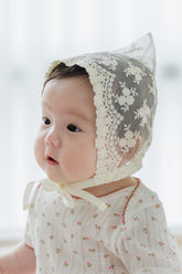 Baby girl wearing Avauma Clare Bonnet in Ivory Race, showing delicate floral lace and satin ribbon detail while held by caregiver